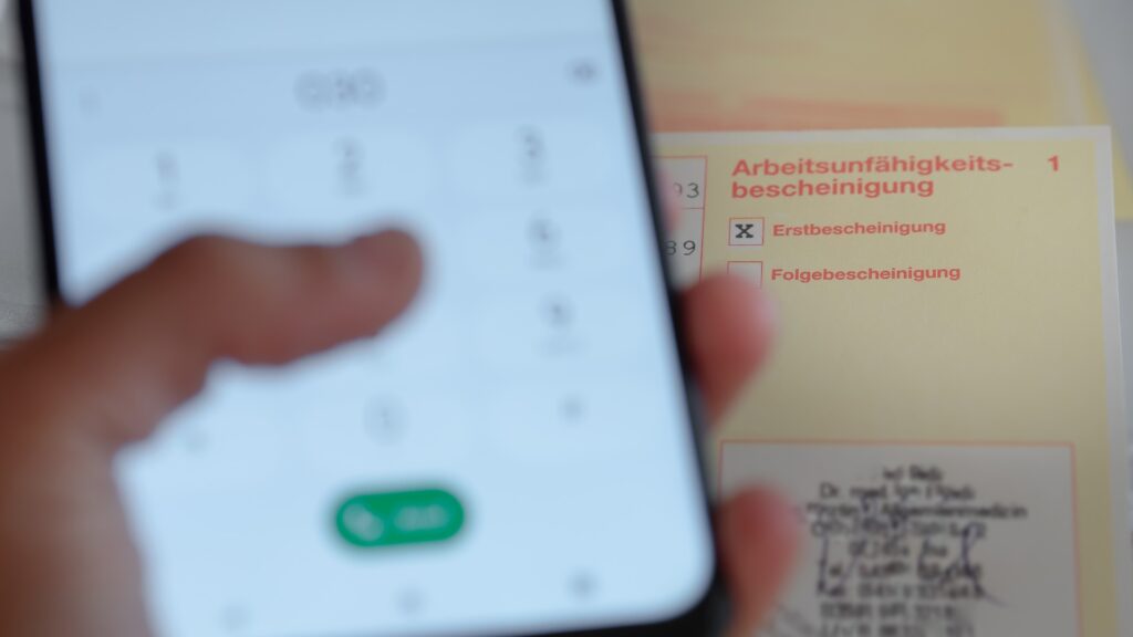 Hand wählt Nummer auf Smartphone und Arbeitsunfähigkeitsbescheinigung